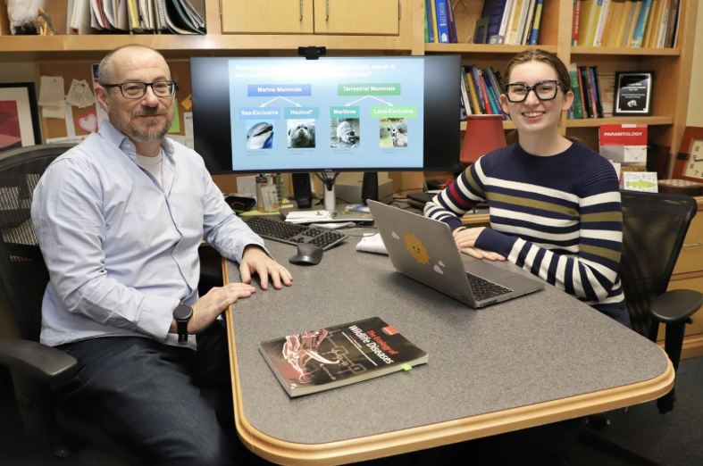 photo of two people sitting across a table, computer screen and books in background