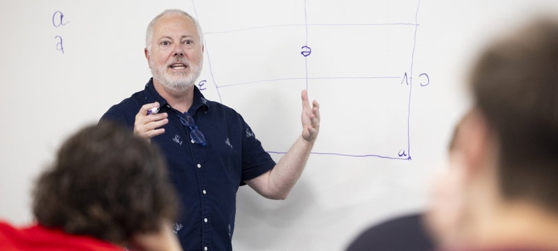 photo of man gesturing, whiteboard in background, students in foreground