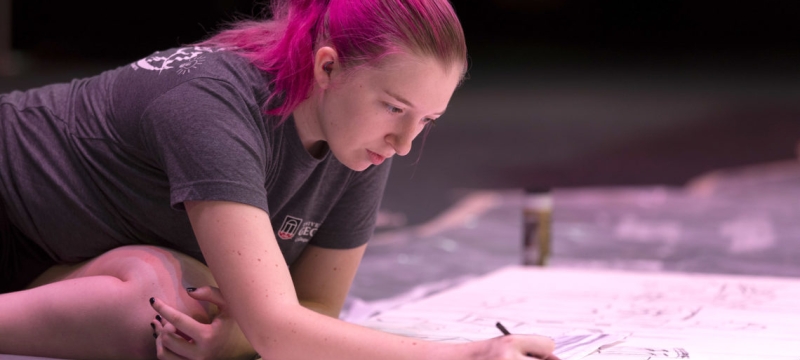 photo of woman leaning over a large drawing surface with pencils