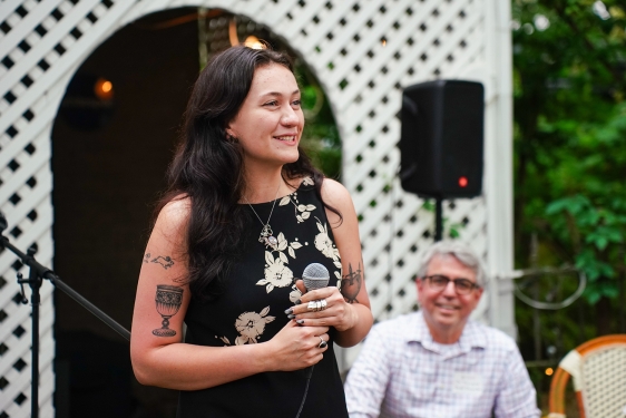 photo of woman with microphone, man in background, outdoors
