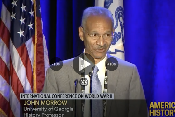 photo of man speaking at microphone, with flags and lower thirds text