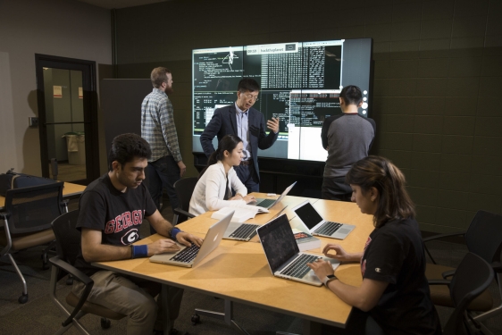 photo of six people in a room with laptop and projection screen, table
