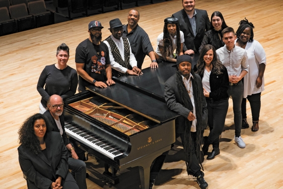 cover photo for magazine with 13 people standing around a grand piano on a concert stage